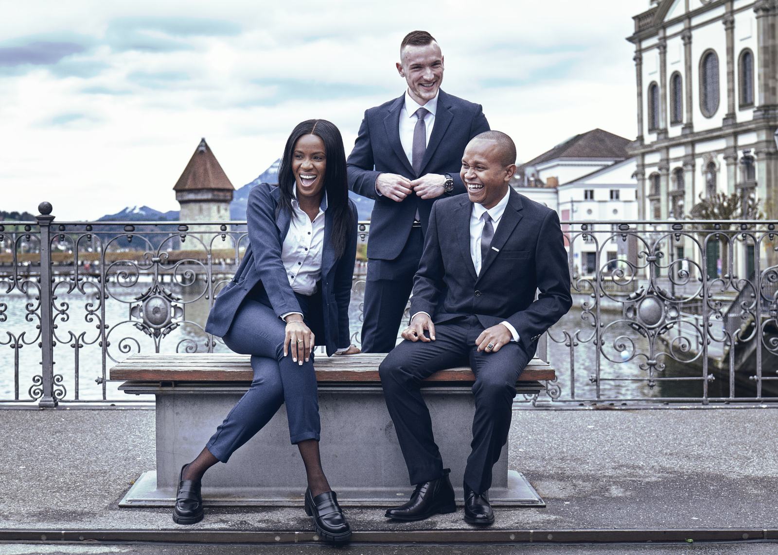 Three BHMS master's students in business dress on the Lucerne waterfront, the Chapel Bridge water tower behind them